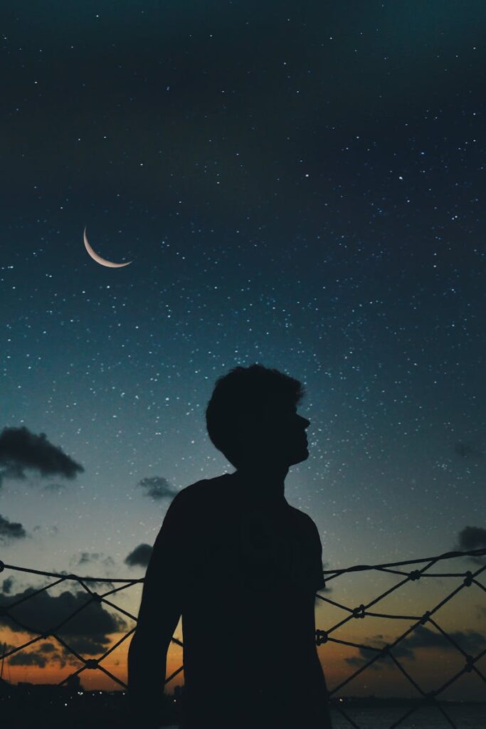 A lone silhouette stands beneath a crescent moon and starry sky, evoking a sense of solitude and wonder.